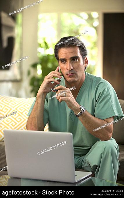Hispanic Male doctor practicing tele-medicine from his home, using cell phone and laptop computer, Listening to patient on video call