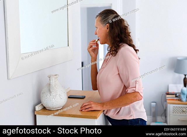 Happy senior caucasian woman in bathroom, looking in mirror, applying lipstick