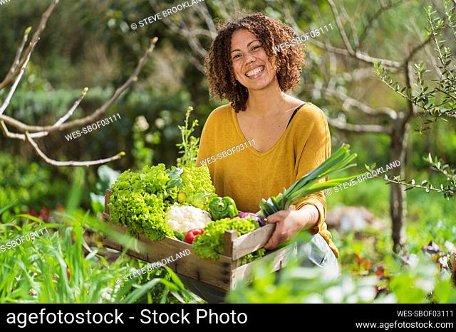 Smiling woman holding a crate with freshly picked organic vegetables in permaculture garden