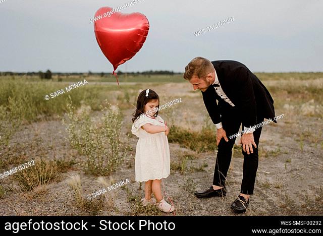 Father talking to sad daughter with heart shape balloon in field against clear sky
