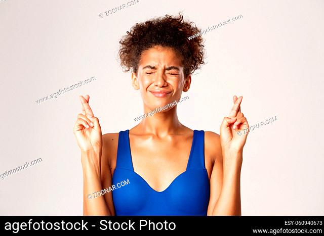Close-up of anxious hopeful girl in fitness clothing, close eyes while making wish or praying, crossing fingers for good luck, white background