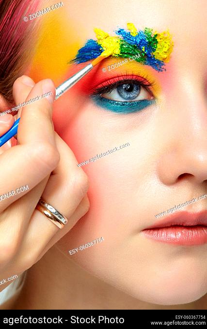Female portrait with unusual face art makeup. Paint on brows. Artist's hand with paintbrush painting beautiful teen girl's brows make-up