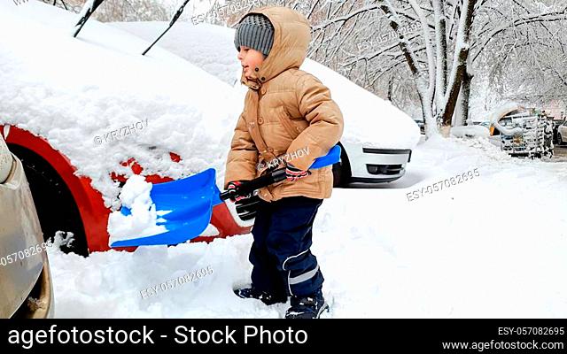 Smiling boy in jacket helping to clean up the snow covered car after blizzard using big blue shovel