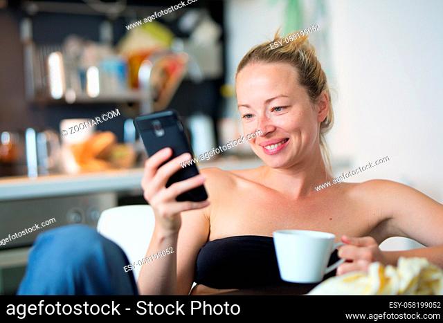 Young smiling cheerful woman indoors at home kitchen using social media on phone for video chatting and staying connected with her loved ones