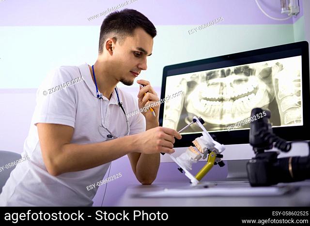 Intent dentist in a white uniform sits on the chair at the table. He looks at an articulator with teeth mould which he holds in the right hand