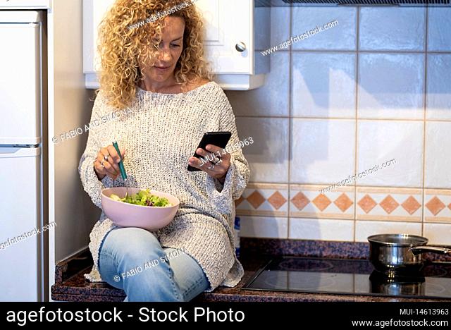 Woman eating healthy salad sitting on the top of the kitchen and home and use diet app on the phone. Health and care free female people indoor leisure activity