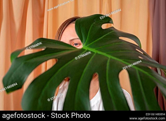 Woman looks through the leaves of the Monstera Deliciosa tropical plant on a fabric curtains background