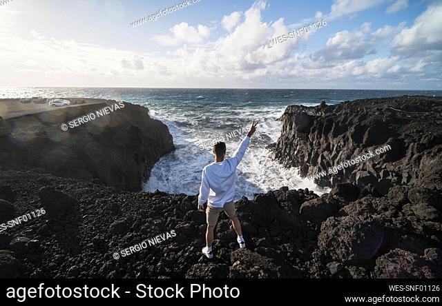 Young male tourist gesturing peace while standing on rock in front of sea at Los Hervideros, Lanzarote, Spain