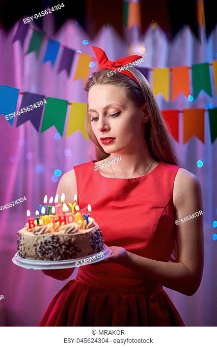 Concept: celebration, birthday. Beautiful young vintage pin up style girl standing in colorful lighted scene holding cake with candles