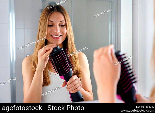 Pleased girl holds round brush hair dryer in her bathroom at home. Young woman holding her salon one-step brush hair dryer and volumizer