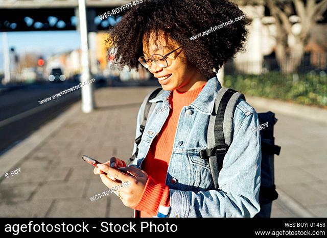 Close-up of smiling young woman with curly hair using mobile phone while standing on sidewalk