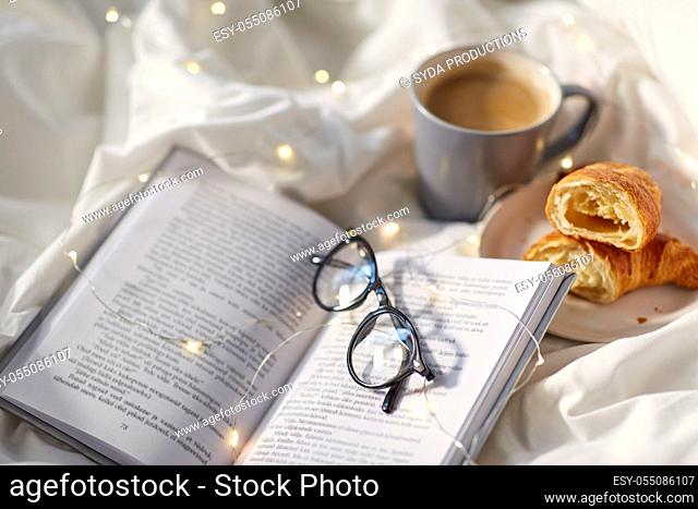 croissants, cup of coffee, book and glasses in bed
