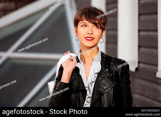 Happy young fashion woman in leather jacket with handbag walking in city street