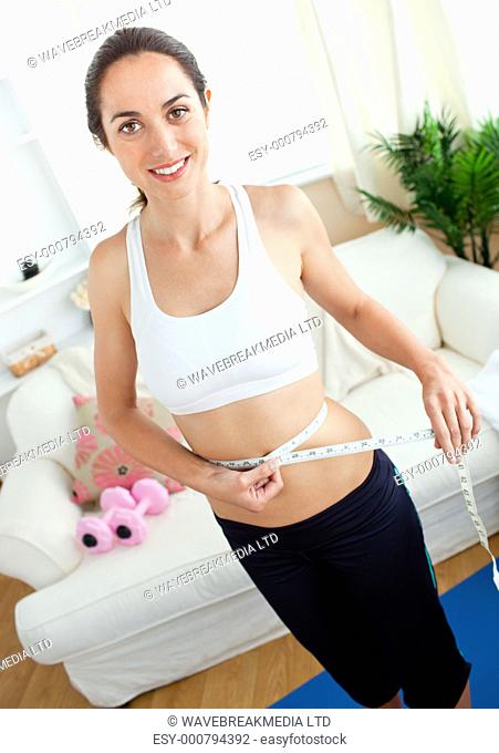 Pretty young woman measuring her waist with a tape in her living-room at home