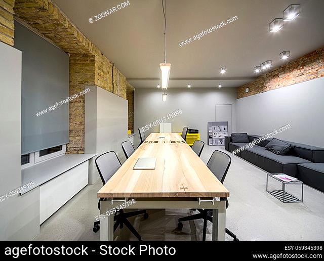 Empty office in a loft style with gray and brick walls. There are glowing lamps, wooden tables with chairs, metal yellow lockers, dark sofas with pillows