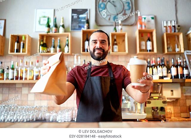 small business, people, takeaway and service concept - happy man or waiter in apron holding coffee cups and paper bag at bar