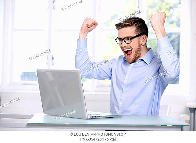 man in a blue shirt, glasses, beard, smiling, window out of focus in the background, mouth opened; hands in the air; sitting, looking at his computer laptop