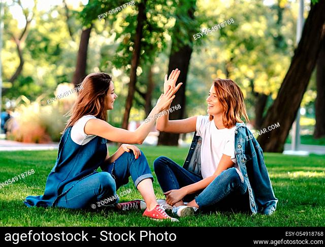 Happy female friends raising hands up giving high five sitting on green grass lawn on sunny day, outdoors in city park