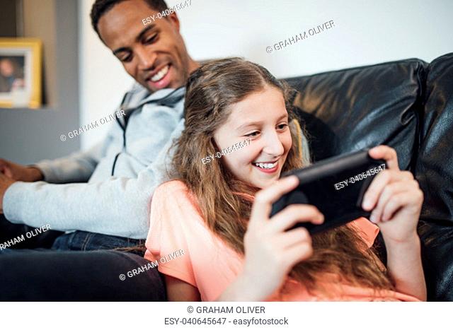 Little girl is playing on a hanheld game console at home and her father is watching her