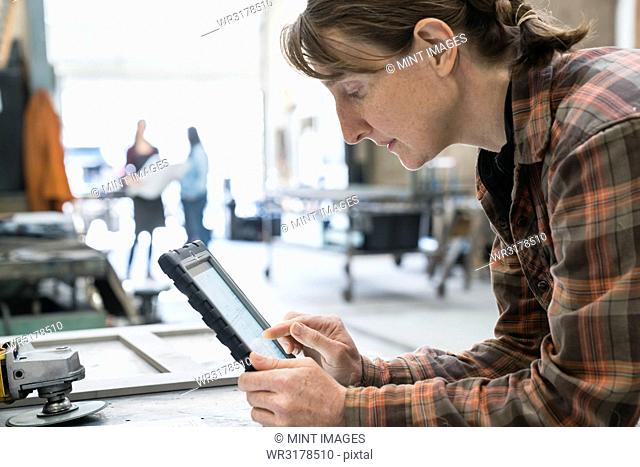 Side view of blond woman wearing checked shirt standing at workbench in metal workshop, holding digital tablet