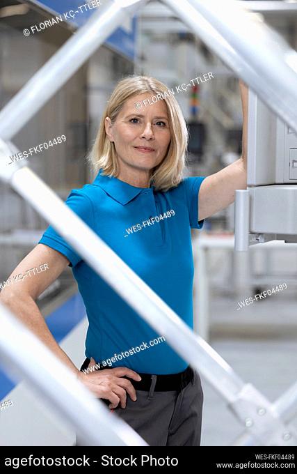 Blond woman with hand on hip standing at machinery seen through railing in factory