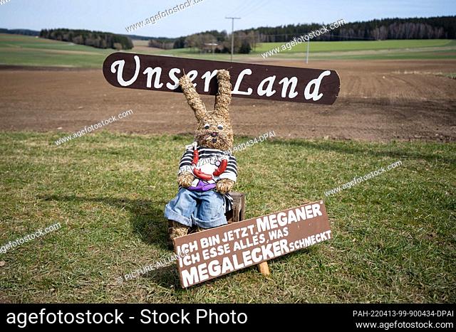 12 April 2022, Bavaria, Silberbach: An Easter bunny made of straw sits behind a sign that reads ""I am now MEGANER. I eat everything that tastes MEGALECKER""