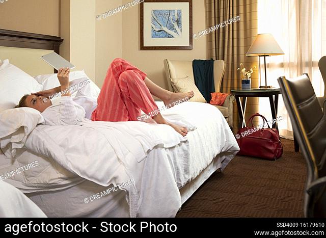Young Caucasian woman with headache wearing bathrobe in a hotel room, lying on bed using tablet device to connect with doctor for a video consultation