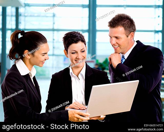 Three young businesspeople standing in lobby, looking at laptop computer screen, smiling