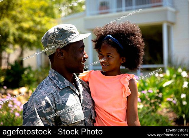 Front view close up of a young adult African American male soldier in the garden outside his home, holding his young daughter