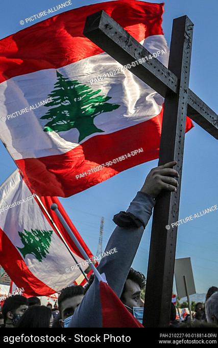 27 February 2021, Lebanon, Bkerki: Anti-government activists wave flags of Lebanon while holding up a cross during a rally at the Maronite Catholic Patriarchate...