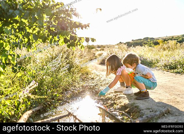 Brother and sister with protective face mask playing at puddle on road during sunny day
