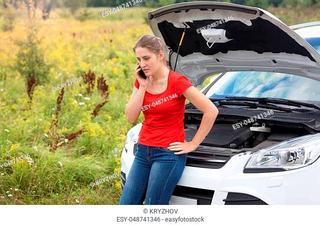 Portrait of sad woman leaning on broken car with open hood and talking by phone