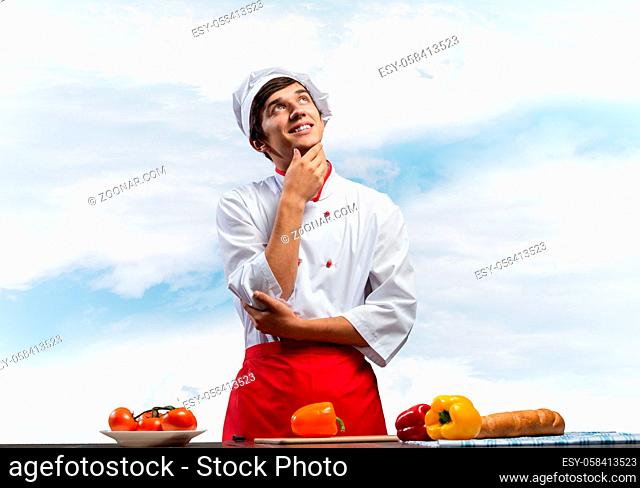 Young male chef standing with folded arms and looking up dreamily. Happy smiling culinary school student in white hat and red apron on blue skyscape background