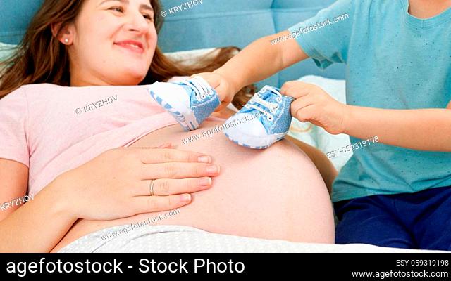 Little toddler boy playing with blue baby boots on big belly of young mother lying on bed. Concept of pregnancy, preparing and expecting child