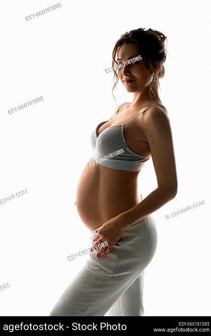 Side view of smiling young pregnant female in white bra and pants looking at camera while standing in light studio