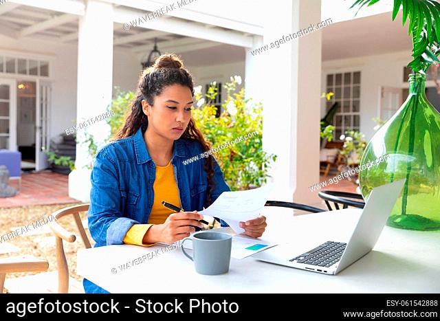 Biracial woman on garden terrace outside house using laptop, drinking coffee and paying bills