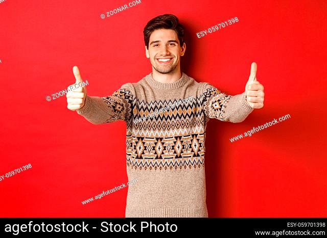 Image of handsome caucasian man in christmas sweater, showing thumbs-up in approval and smiling, wishing happy holidays, standing over red background