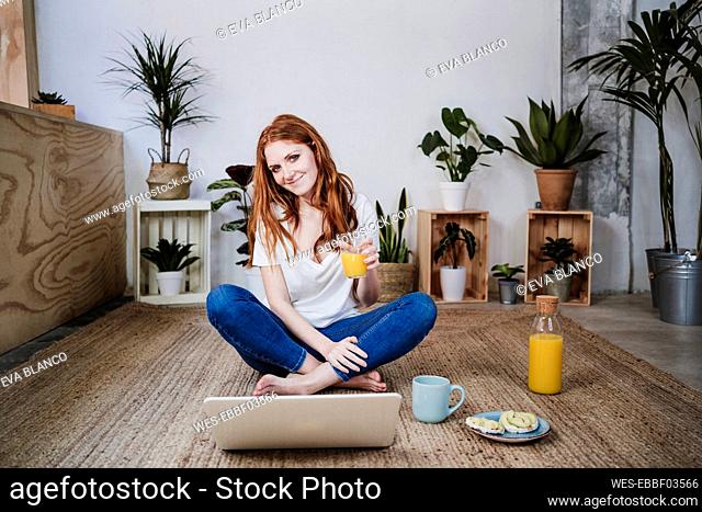 Smiling young woman sitting cross legged while having juice in front of laptop on floor at home