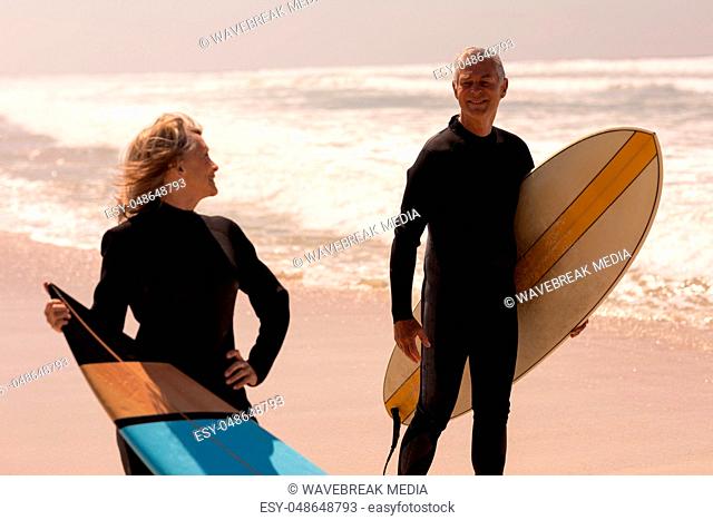 Senior couple with surfboard standing on beach and looking each other