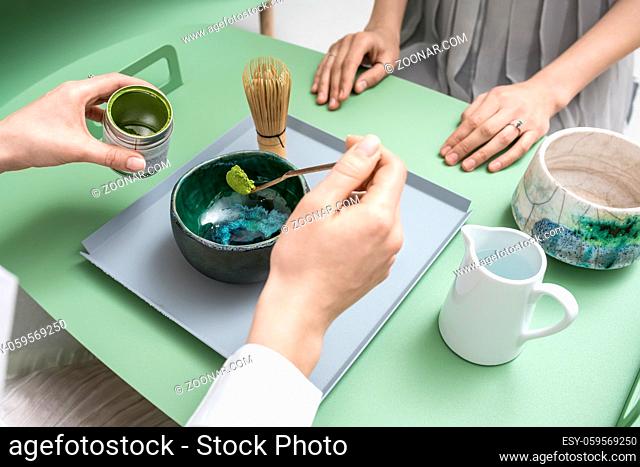 People preparing a chinese matcha green tea on the green metal table indoors. One woman measures tea powder with a wooden stick over the bowl