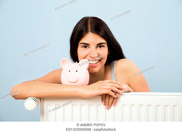 Young Happy Woman With Piggybank On Radiator At Home