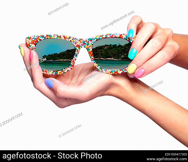 Woman is holding fancy sunglasses with sea beach reflection. Female fingers with bright green, yellow, pink and blue nails manicure
