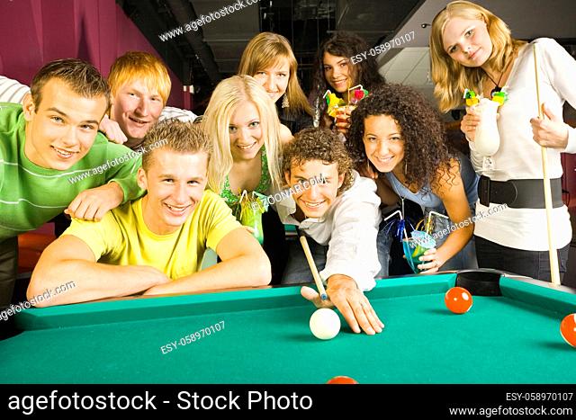 Large group of teenagers standing at pool table. Smiling and looking at camera. One person is playing billard, some of them holding drinks