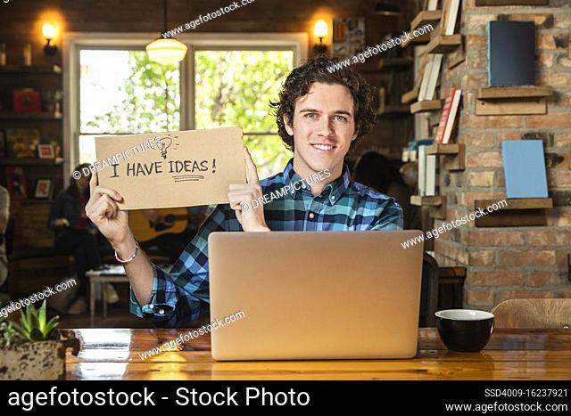 Portrait of young man sitting at table in coffee shop bookstore with laptop computer, holding hand made sign that reads ?I have ideas