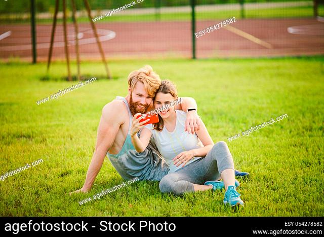 Sport and technology. Young in love heterosexual Caucasian couple resting after workout outdoors in park on lawn, green grass sitting in embrace and making...