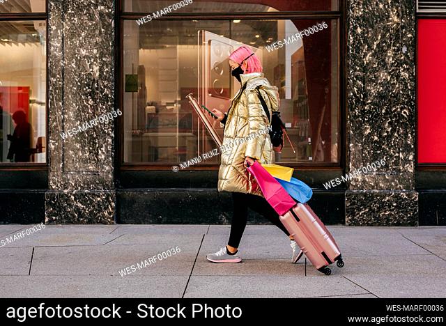 Young woman wearing face mask using mobile phone while walking with shopping bags and luggage on footpath