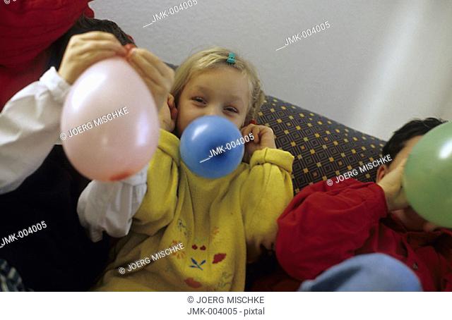 Three children, boys and a girl, 5-10 years old, sitting on a sofa, inflating balloons
