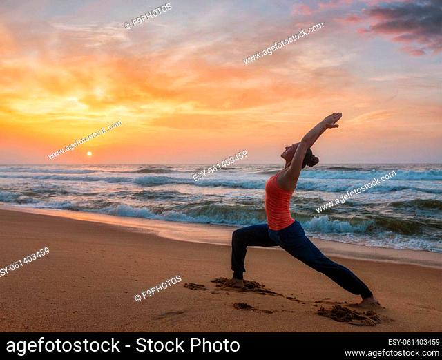 Woman doing Hatha yoga asana Virabhadrasana 1 Warrior Pose outdoors on ocean beach on sunset. Kerala, India