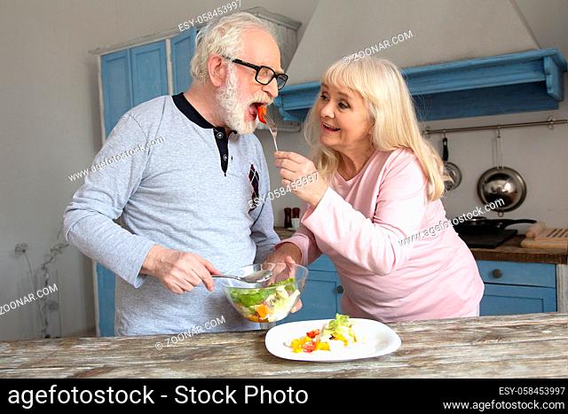 Couple of cute aged people preparing meal. Cute elderly woman dressed in light pink feeding her husband with salad for lunch