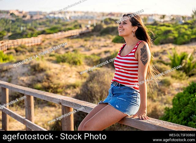 Thoughtful teenage girl sitting on wooden railing at beach during sunny day
